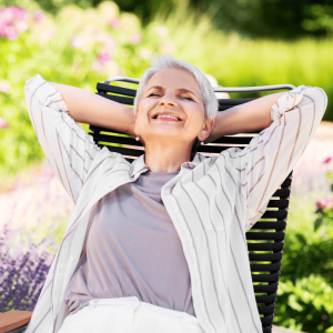 Older Lady sitting relaxing on a chair in her garden enjoying the Benefits of Artificial Grass. She has a purple top on with a white stripy shirt and white trousers. She is in her garden which has artificial grass as well as some well looked after flowers.